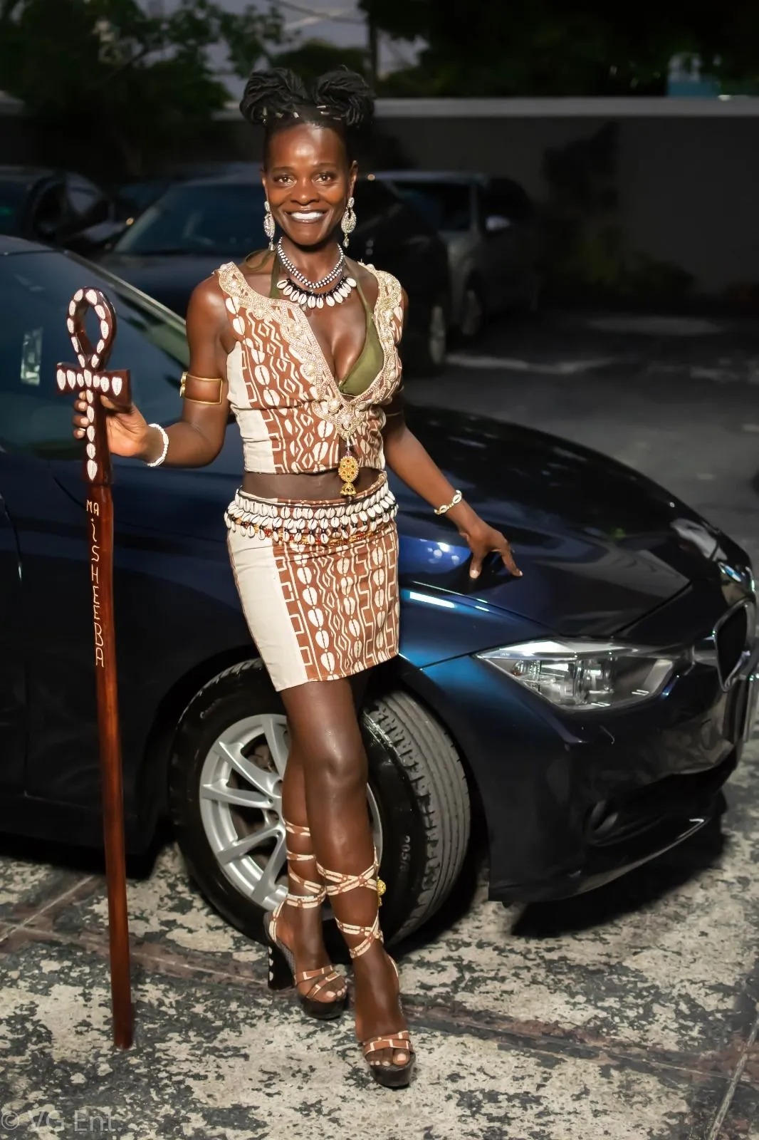 Confident woman wearing a traditional African-inspired outfit, holding a carved staff and standing beside a car at night.