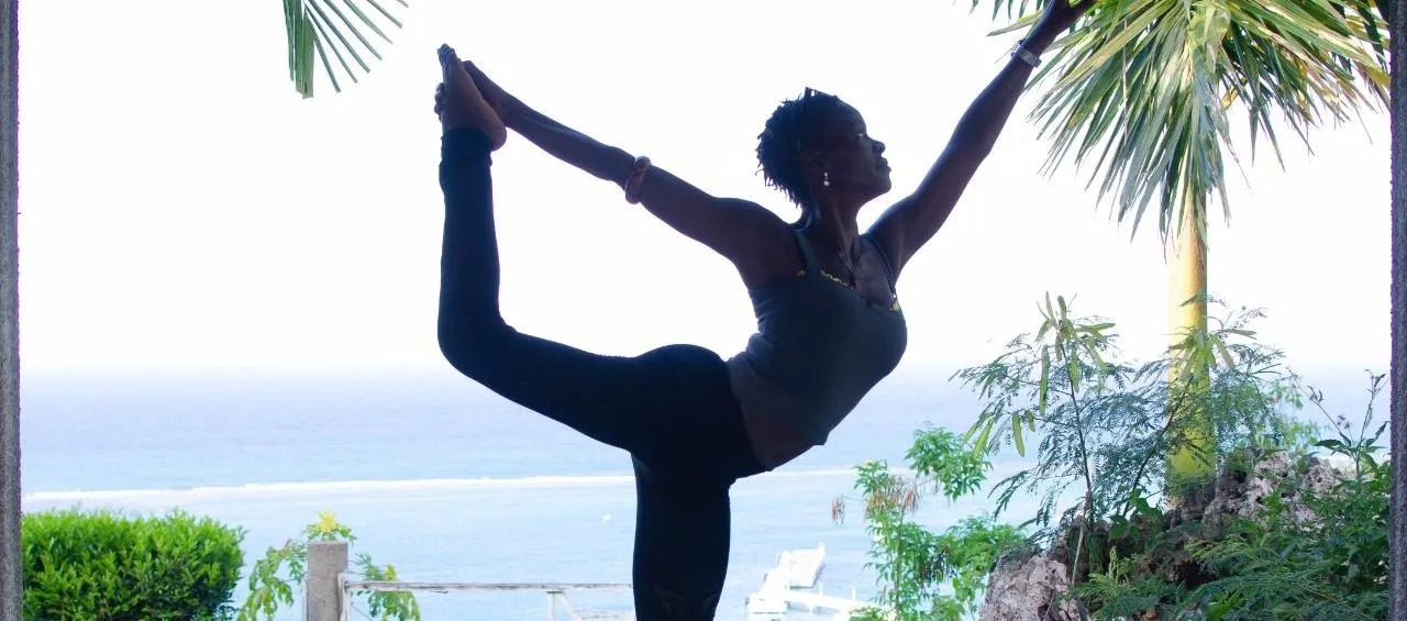 Woman meditating on a beach at sunset with a group practicing yoga in the background.