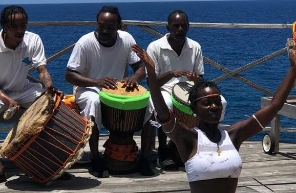 Group of musicians playing traditional drums while a woman raises her arms in expressive movement by the ocean.